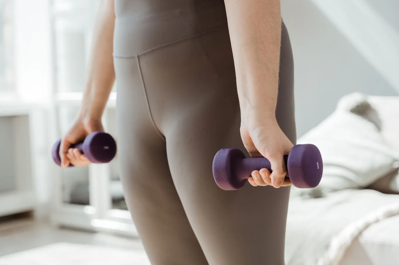 Person holding a pair of purple dumbbells while standing in a home bedroom