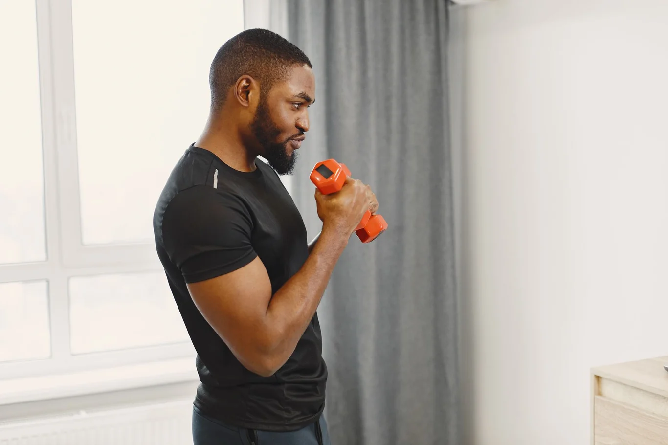 Man performing a bicep curl with an orange dumbbell beside a window