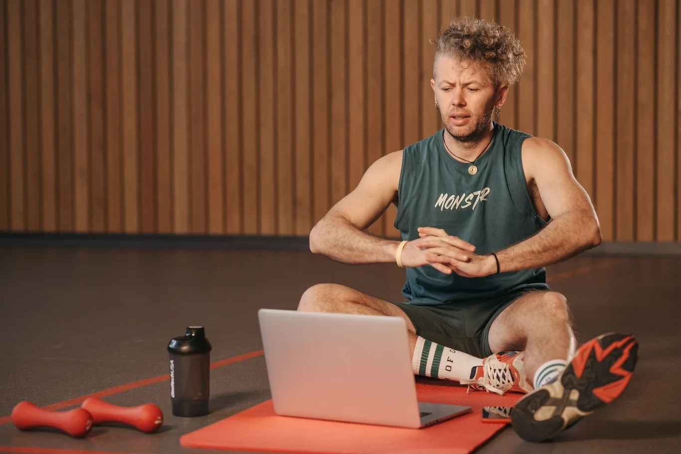 Man sitting on a mat with dumbbells and a resistance band watching a laptop screen