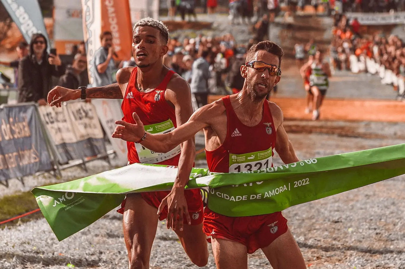 Two runners in red uniforms breaking through a green finish line tape at a road race with spectators in the background