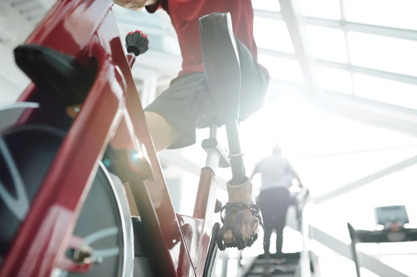 Person cycling on a red exercise bike in a bright gym with a treadmill runner visible in the background