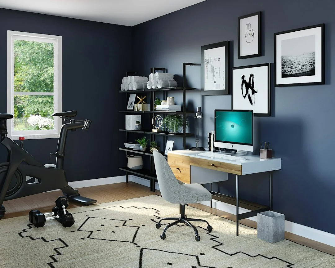 Stationary spin bike beside a home office desk with dark walls and shelving
