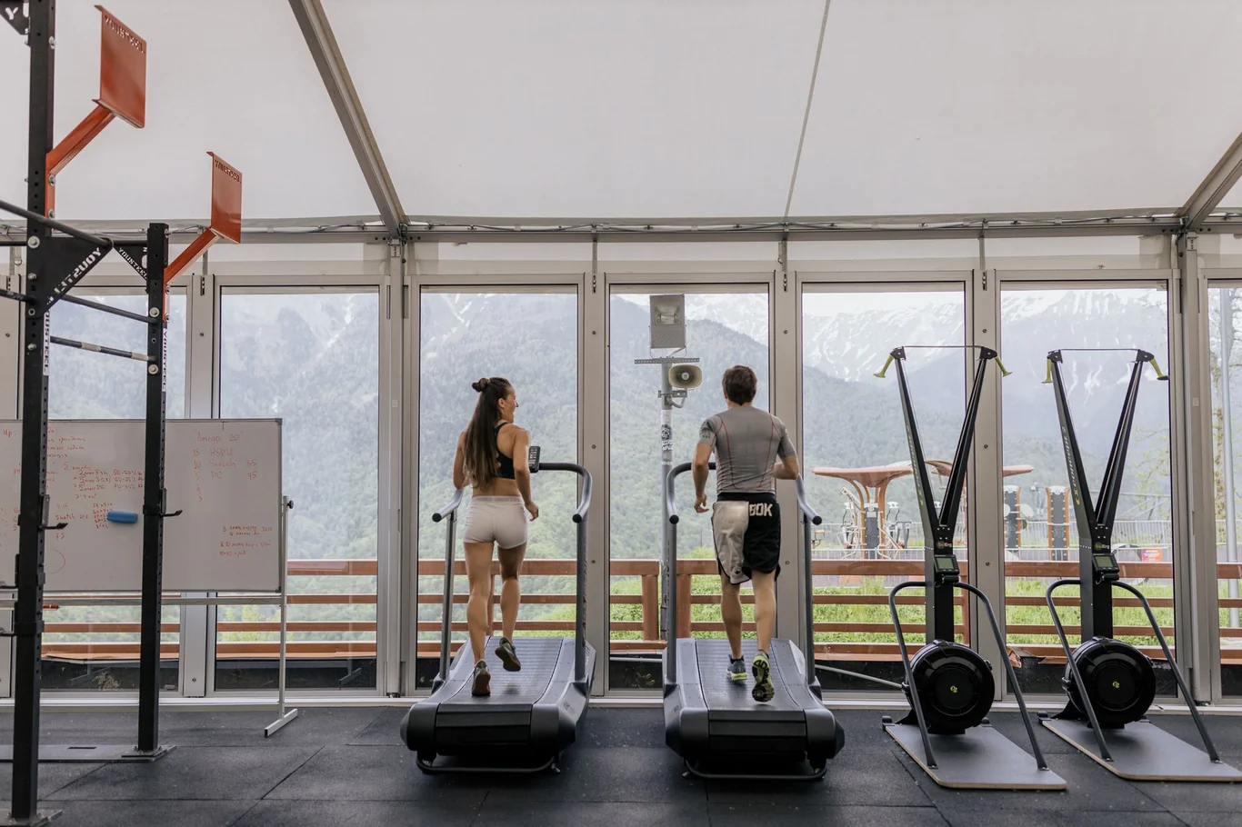 Two people running on treadmills in a bright gym with mountain views through floor-to-ceiling windows