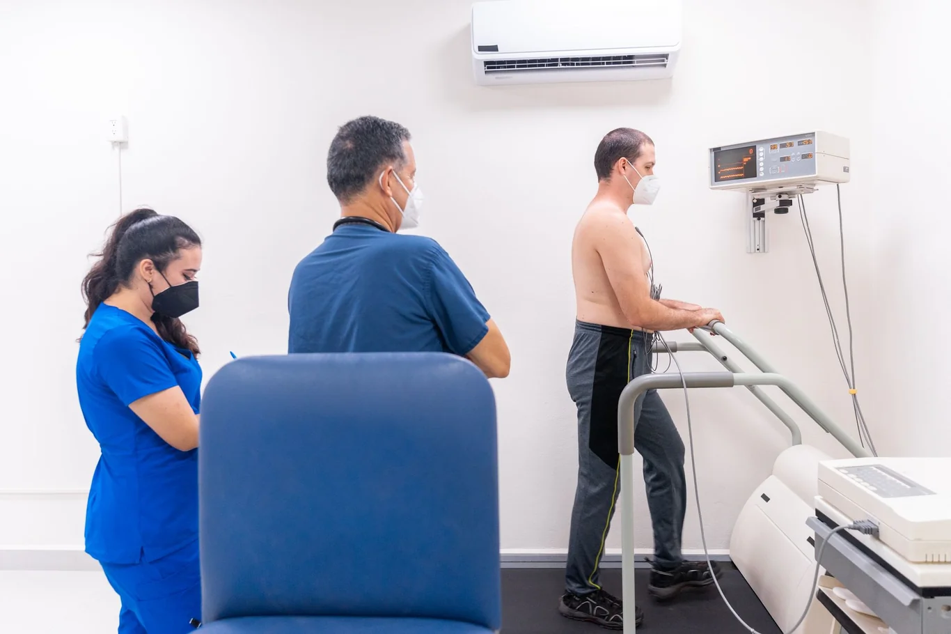 Shirtless man walking on a treadmill during a clinical stress test as two masked health professionals monitor