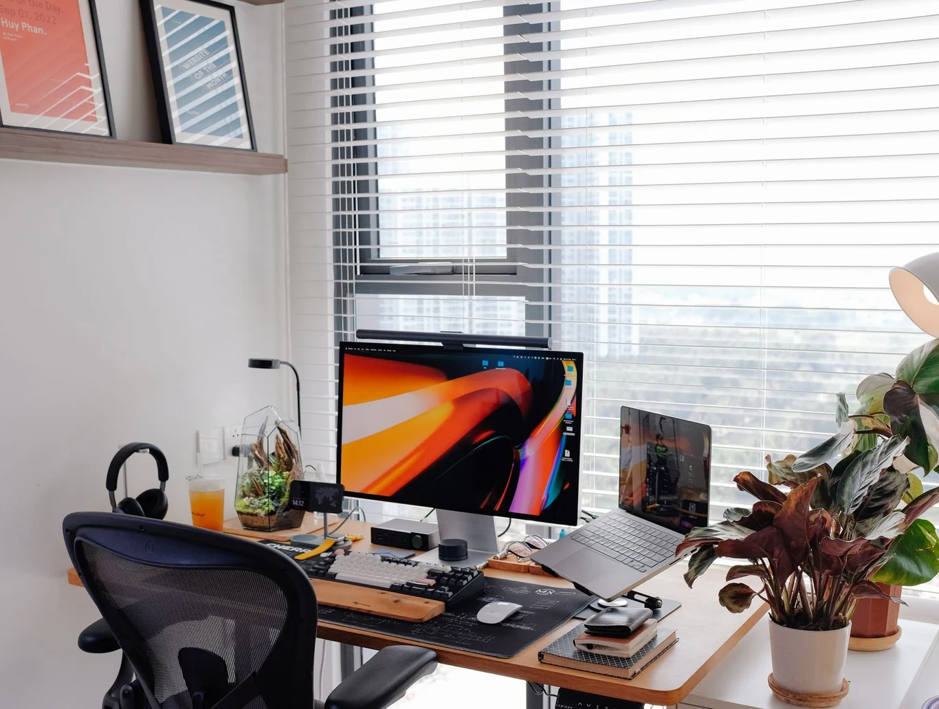 Home office desk setup with a monitor, laptop, headphones, and plants near a window