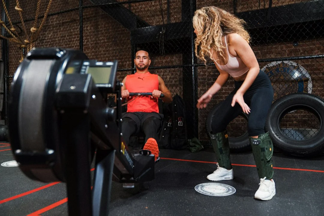 Man rowing on an exercise machine while a woman trains with weights in a gym