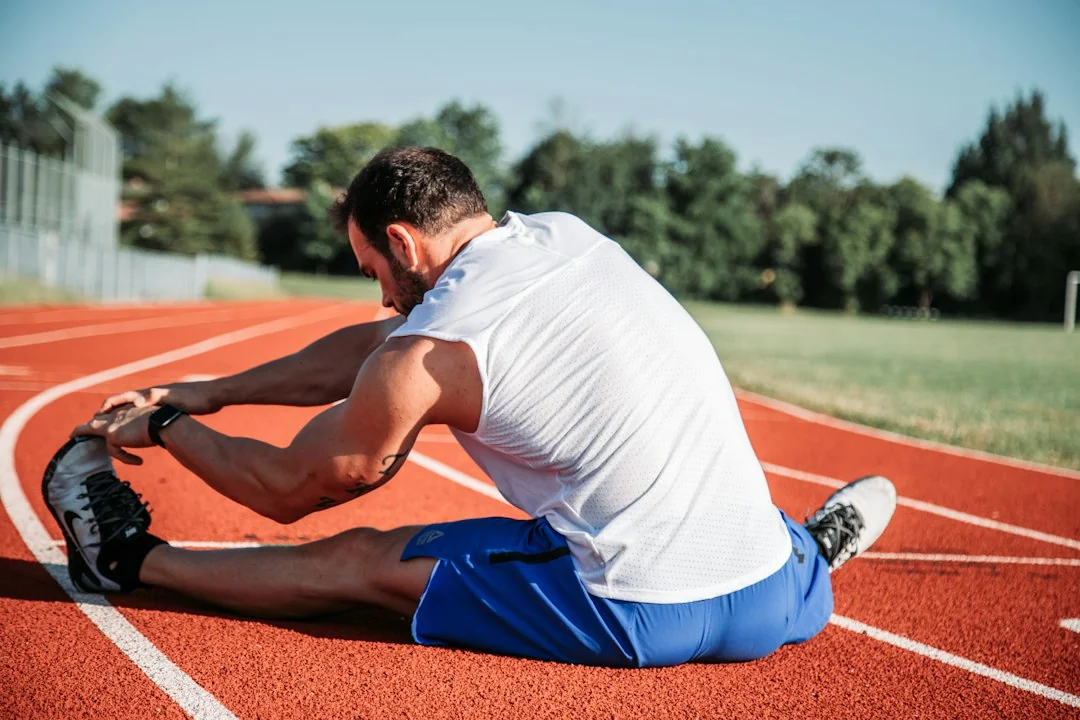 Athlete stretching forward on a red running track in natural sunlight with trees in the background