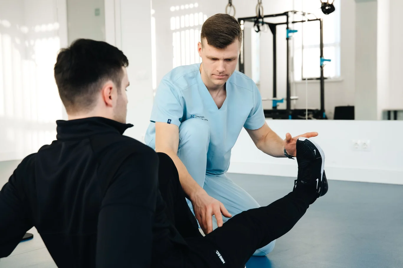 Physical therapist in blue scrubs guiding a patient through a leg exercise in a rehabilitation gym