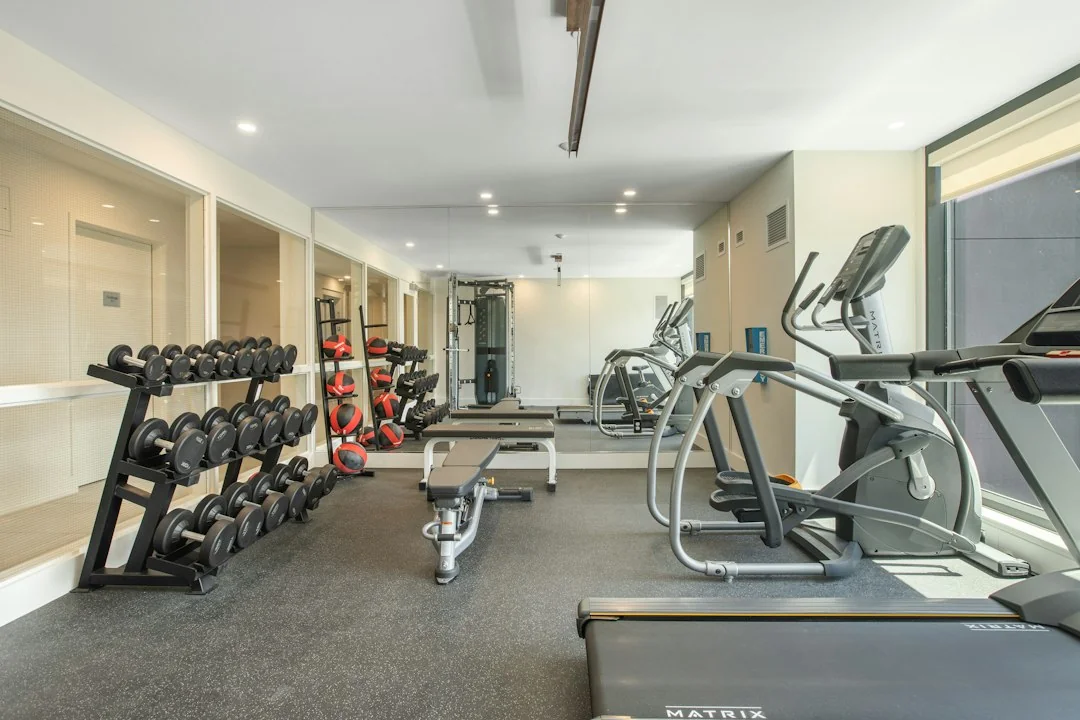 Modern gym floor with dumbbells rack, treadmills, and elliptical machine under bright overhead lighting