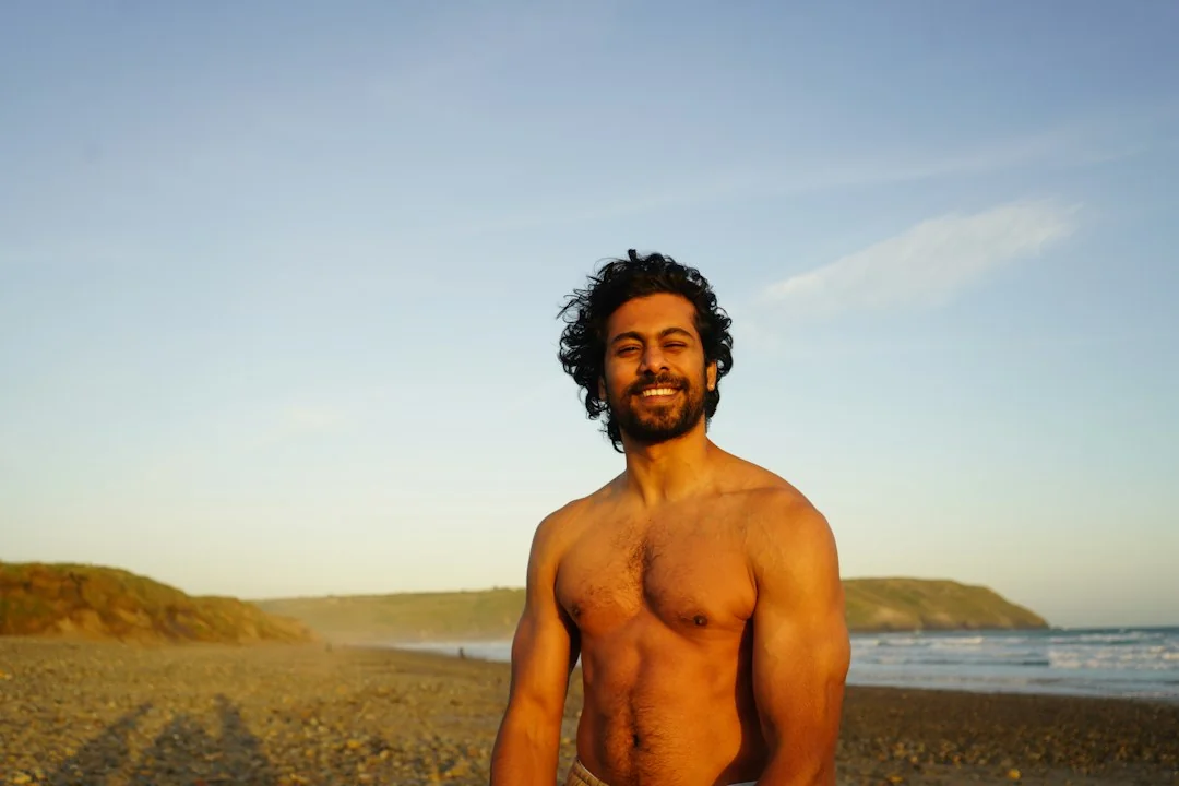 Lean athletic man standing shirtless on a sandy beach at golden hour, smiling with ocean in background