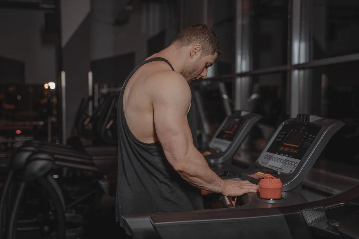 Athletic man at a treadmill in a gym holding a water bottle, weight racks visible behind him
