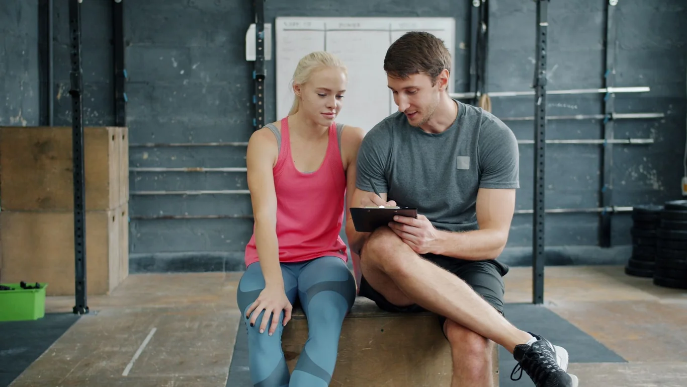 Personal trainer reviewing a fitness plan on a clipboard with a female client seated in a gym