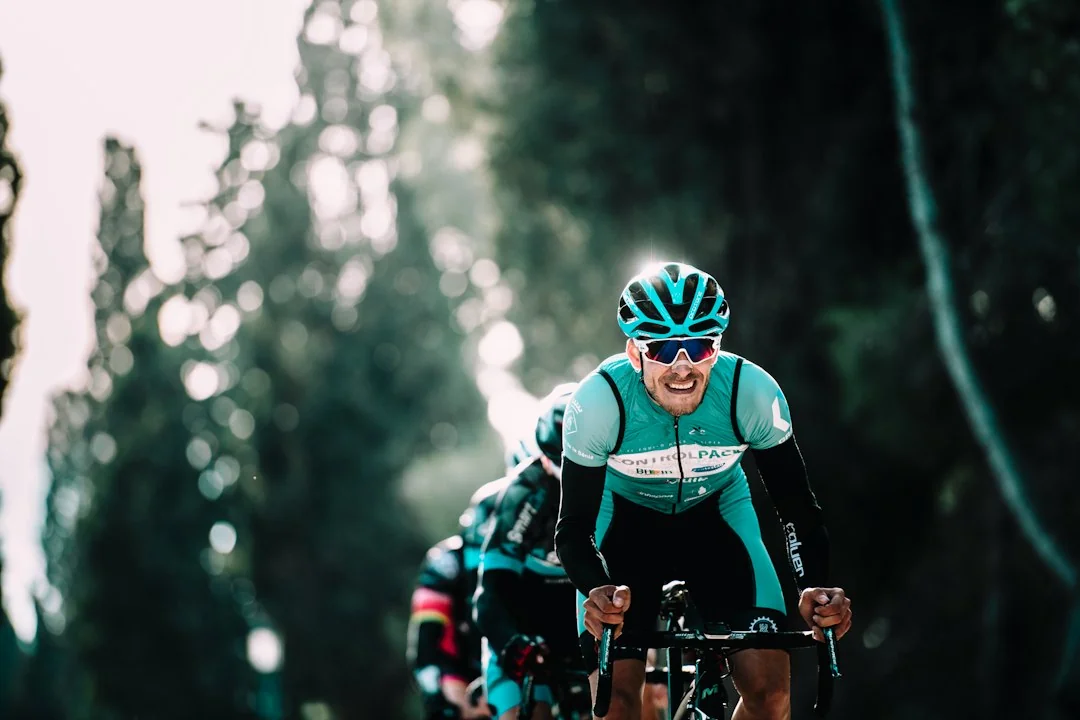 Road cyclist in teal kit leading a group ride through a tree-lined course in natural sunlight