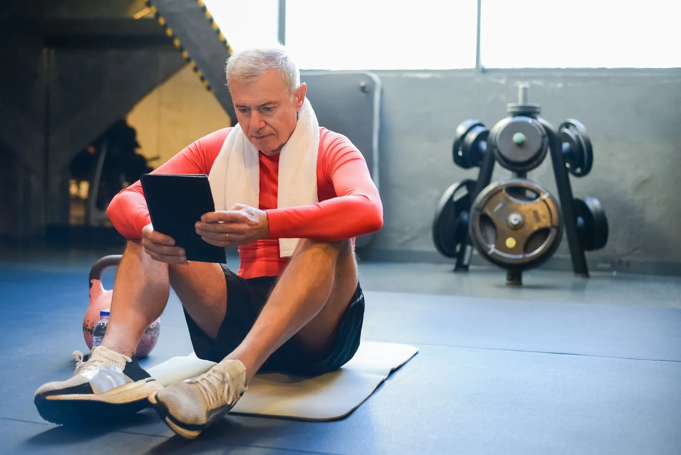 Man in red workout shirt sitting on gym floor reviewing a tablet after exercise