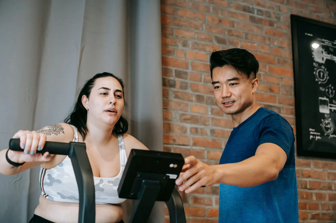 Woman riding a stationary cycling bike with a screen while a trainer points at the display