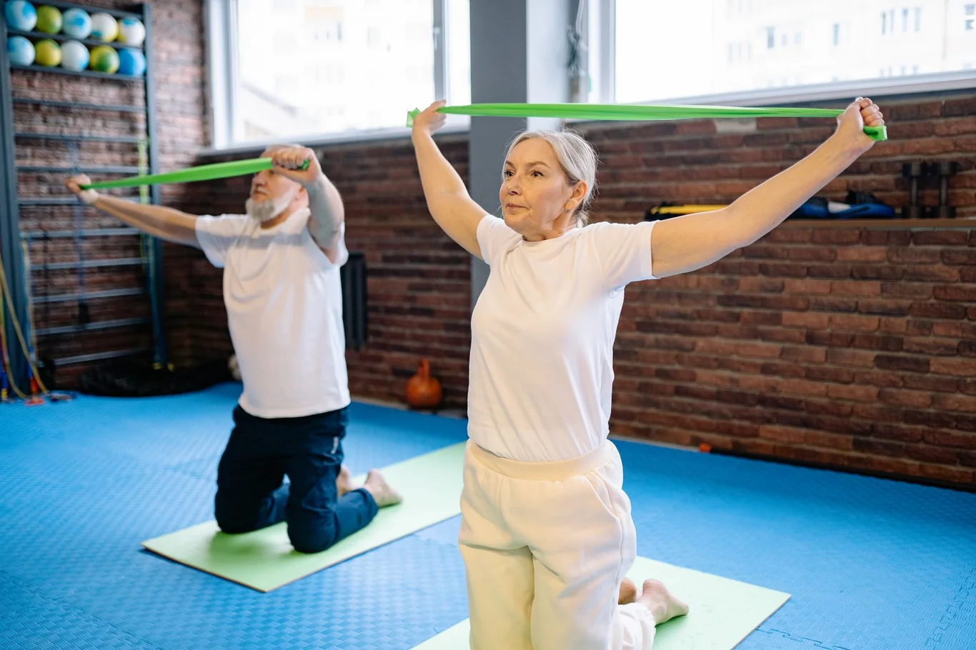 Senior woman kneeling on a mat and stretching a resistance band overhead in a gym