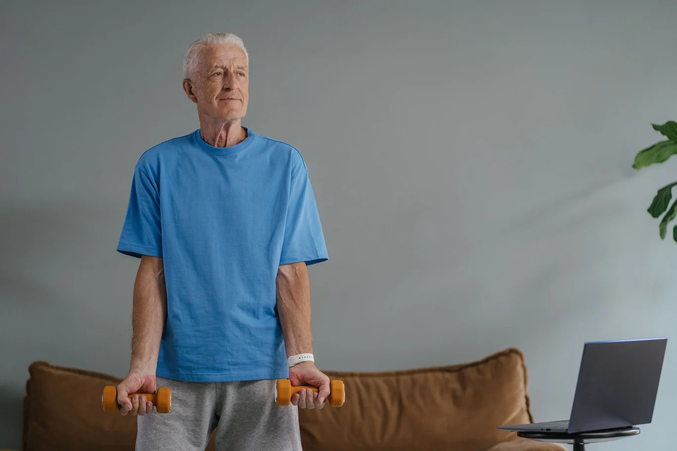 Elderly man holding light orange dumbbells while standing in a home living room