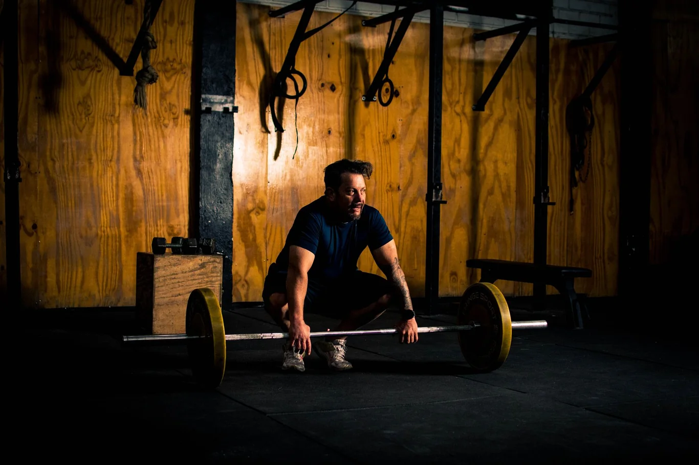 Man crouching before a loaded barbell in a dark gym, preparing to lift