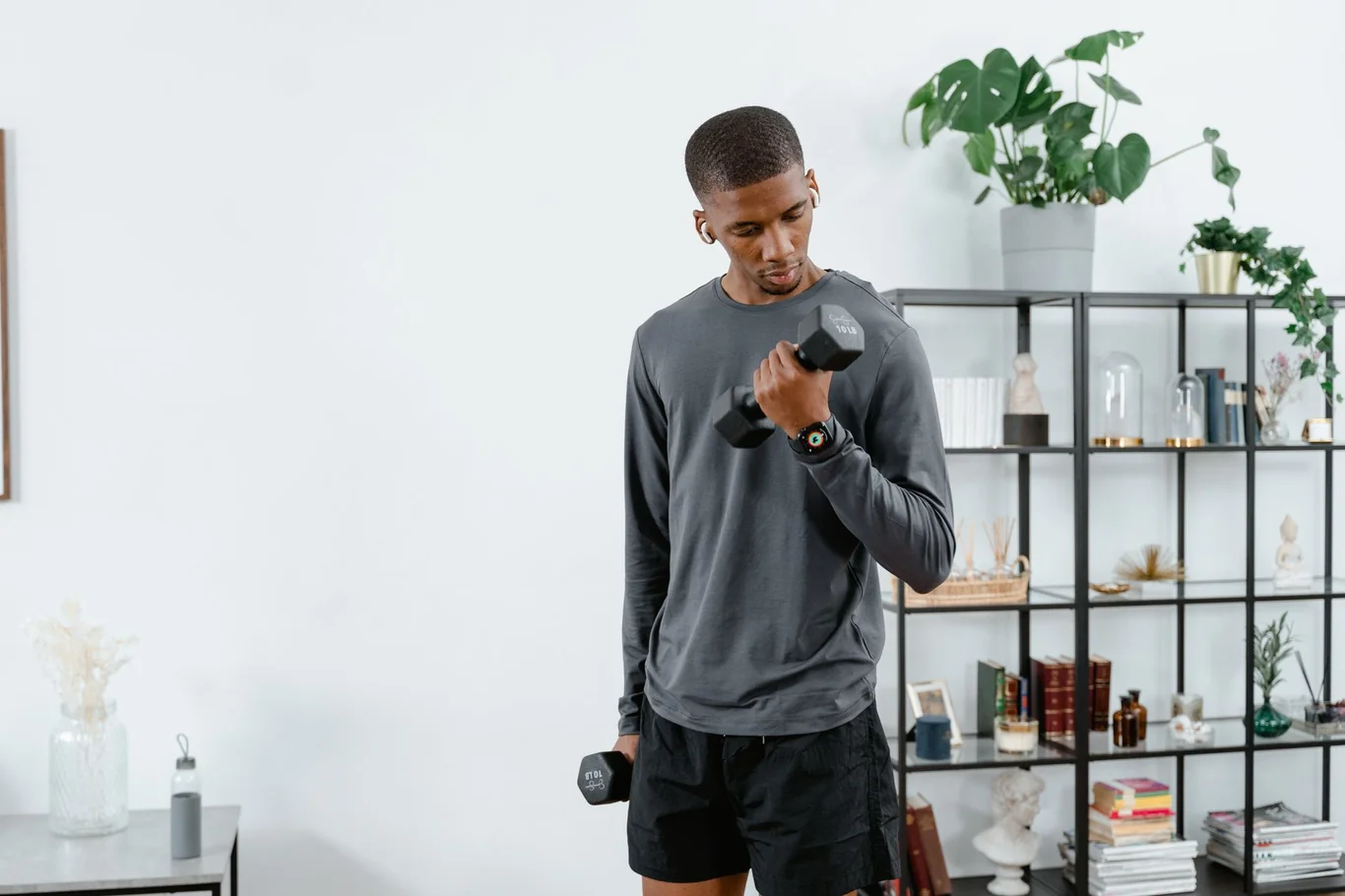 Man performing a dumbbell bicep curl in a bright, compact apartment with a shelving unit behind him