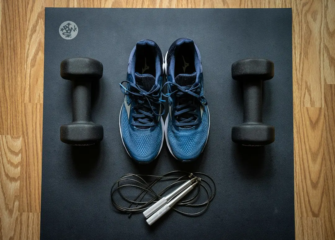 Flat-lay of dumbbells, blue running shoes, and a jump rope on a black exercise mat over wood floor