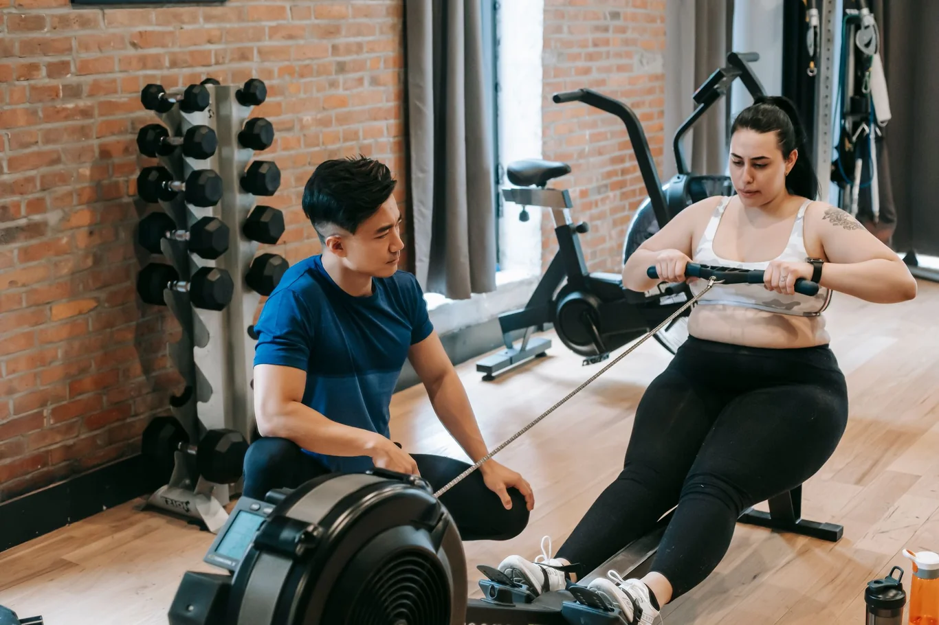 Woman rowing on an ergometer in a gym while a trainer observes