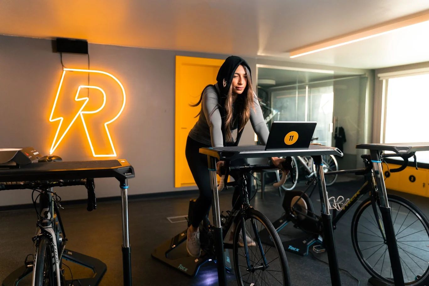 Woman riding a stationary bike in a gym studio while following a live workout class on a laptop
