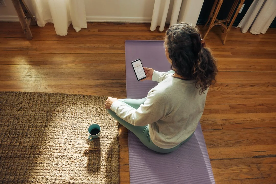 Woman sitting on a yoga mat at home browsing a fitness app on her phone