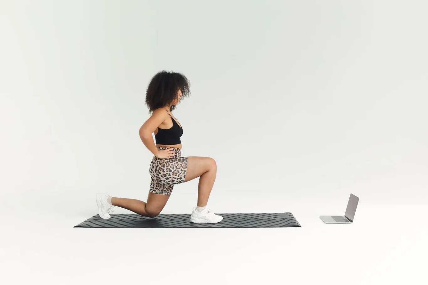 Woman doing a lunge on a yoga mat with a laptop open beside her