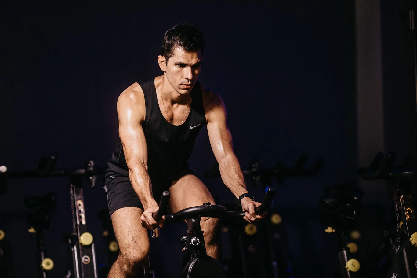 Muscular man riding a spin bike at high intensity in a dark indoor cycling studio
