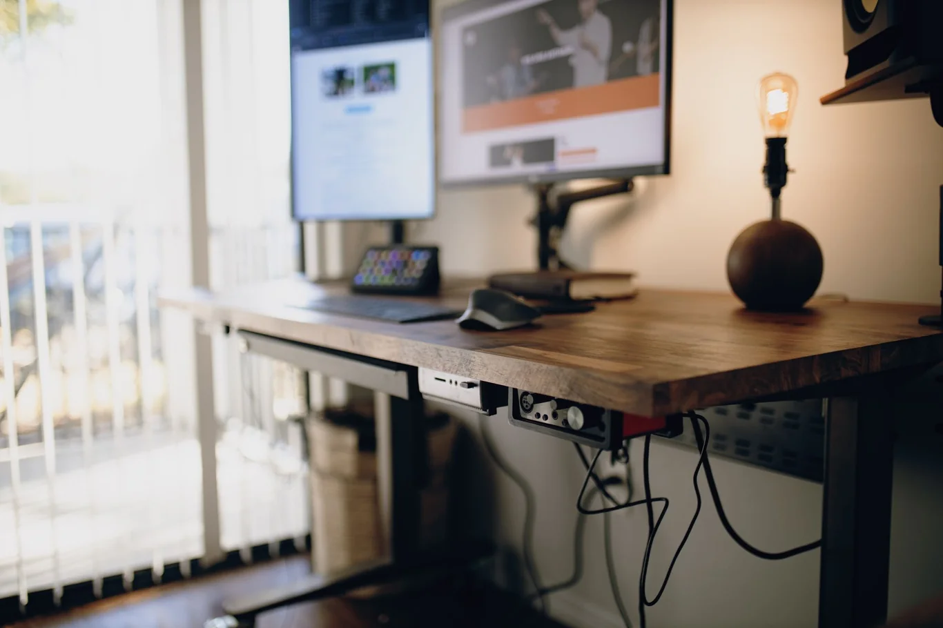 Wooden standing desk with dual monitors and a warm lamp in a home office