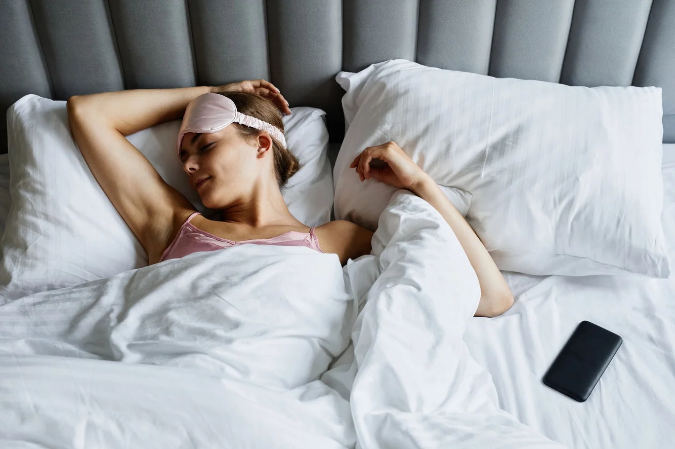 Woman sleeping in bed with a pink eye mask and a smartphone beside her