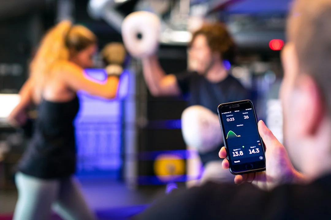 Person checking a fitness tracking app on a smartphone while boxers train in the background