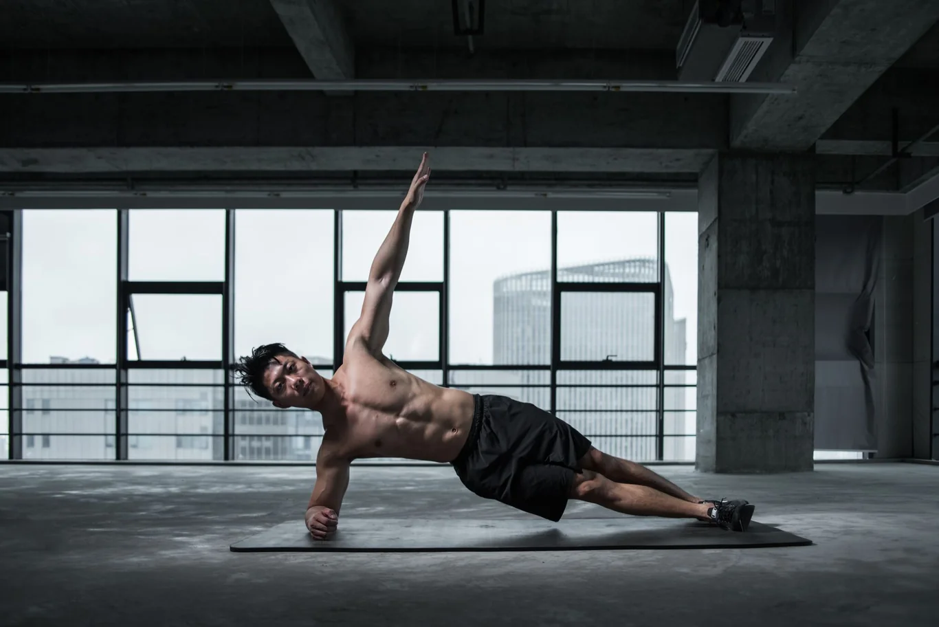 Man performing a side plank on a gym floor with city skyline visible through windows